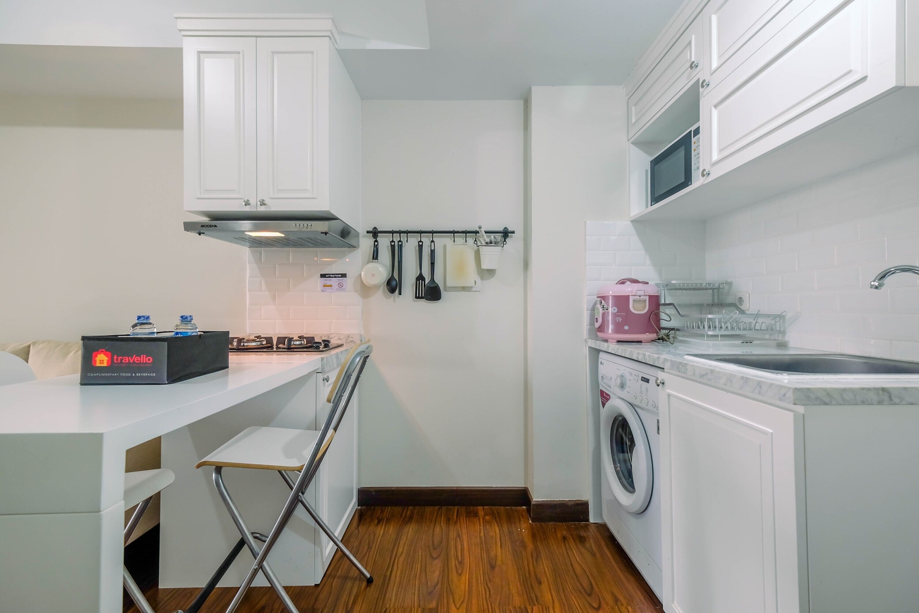 A compact kitchen with white cabinets, a microwave, a washing machine, and a small bar stool.