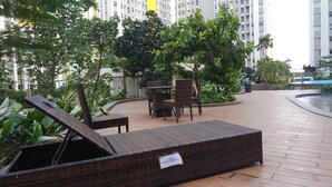 Juego de sillón y mesita de mimbre en un espacio exterior con piso de baldosas, rodeado de follaje y una piscina.