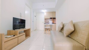 Una sala moderna con un televisor de pantalla plana, mueble de entretenimiento de madera, sofá beige y una pequeña área de cocina con gabinetes de madera y una encimera blanca.