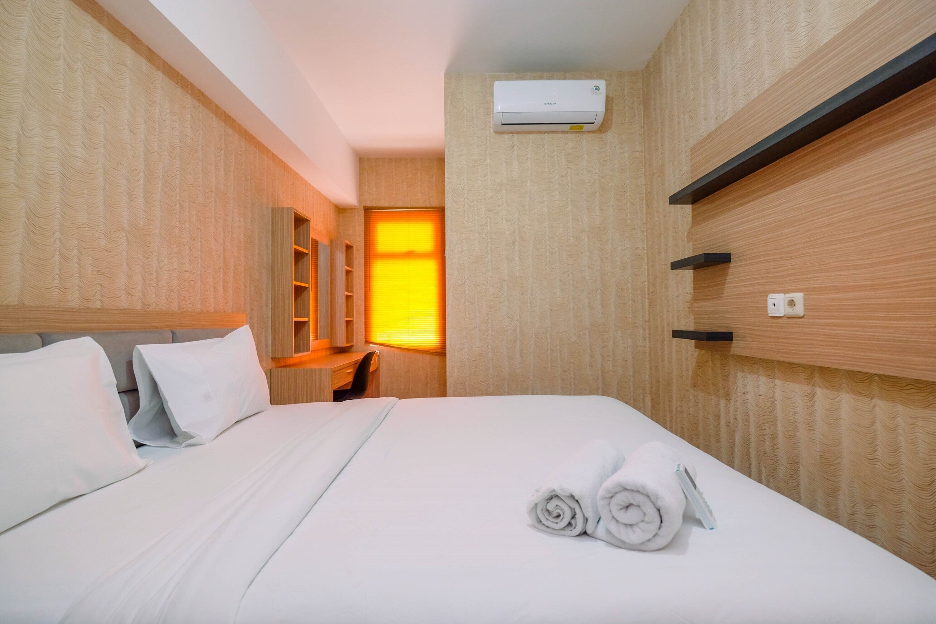 A hotel room with a bed, a desk, a chair, a mirror, and an air conditioning unit.