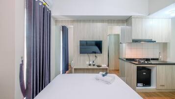 A hotel room with a bed, a TV mounted on the wall, a small kitchen area with a sink and stove, and a bathroom visible through a curtain.