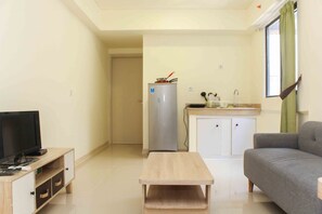 A living room with a sofa, a coffee table, a TV, a kitchen area with a fridge, and a window with curtains. - Best Choice and High Floor 1BR at Meikarta Apartment (Cikarang)
