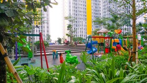 Parque infantil com escorregadores e estruturas de escalada, cercado por vegetação e edifícios altos.