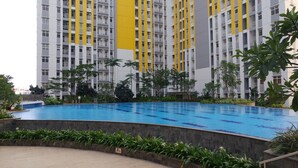 A swimming pool surrounded by greenery and a high-rise building with yellow accents. - Relaxing 2BR Apartment at The Springlake Summarecon (Bekasi)