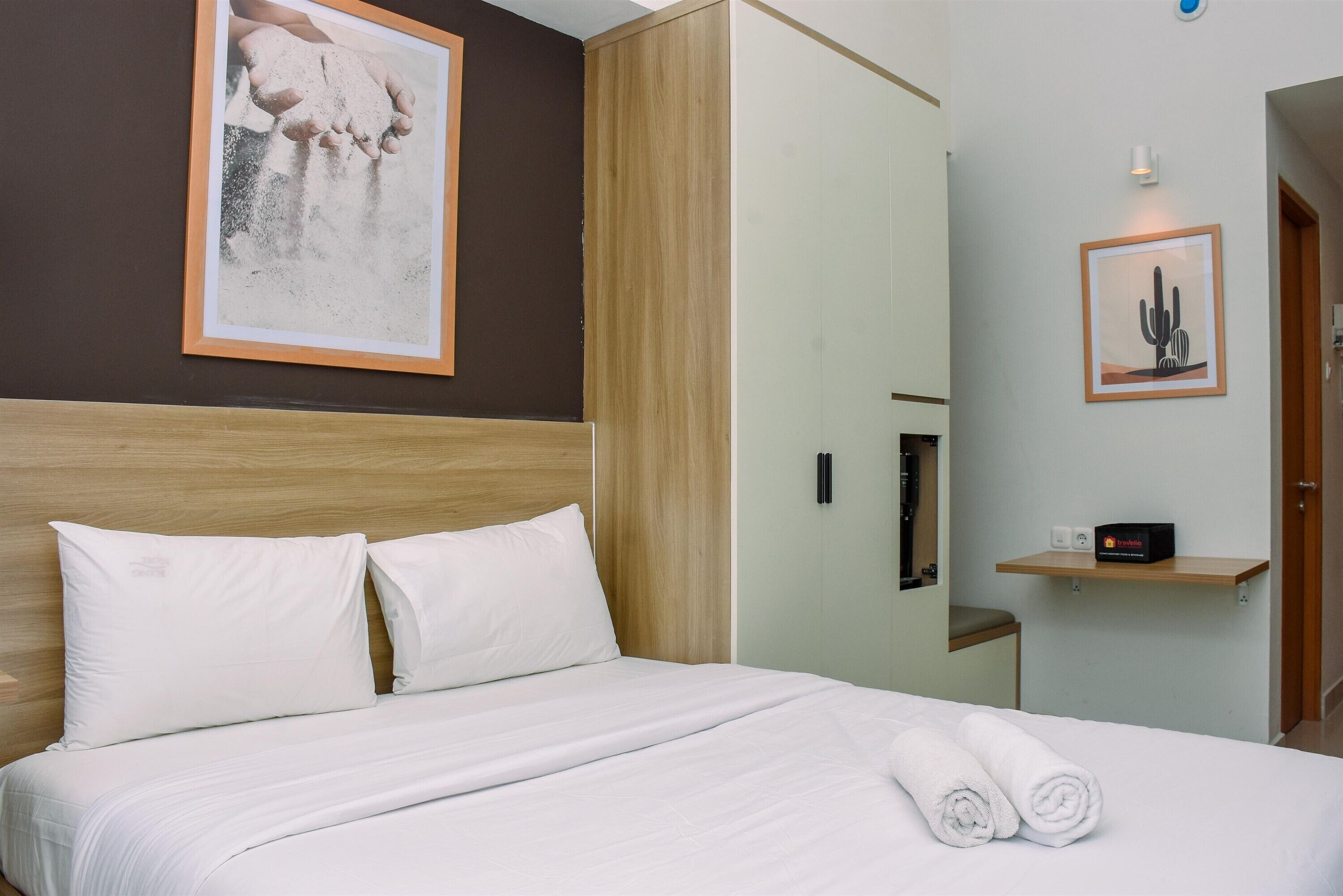 A neatly made bed with white linens and two pillows, a framed picture of hands holding sand, a wooden headboard, a wooden shelf with a cactus picture, and a door to the right.