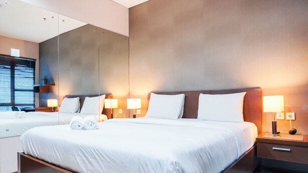 A modern hotel room with a large bed, two bedside tables with lamps, a mirror, and a window with blinds.