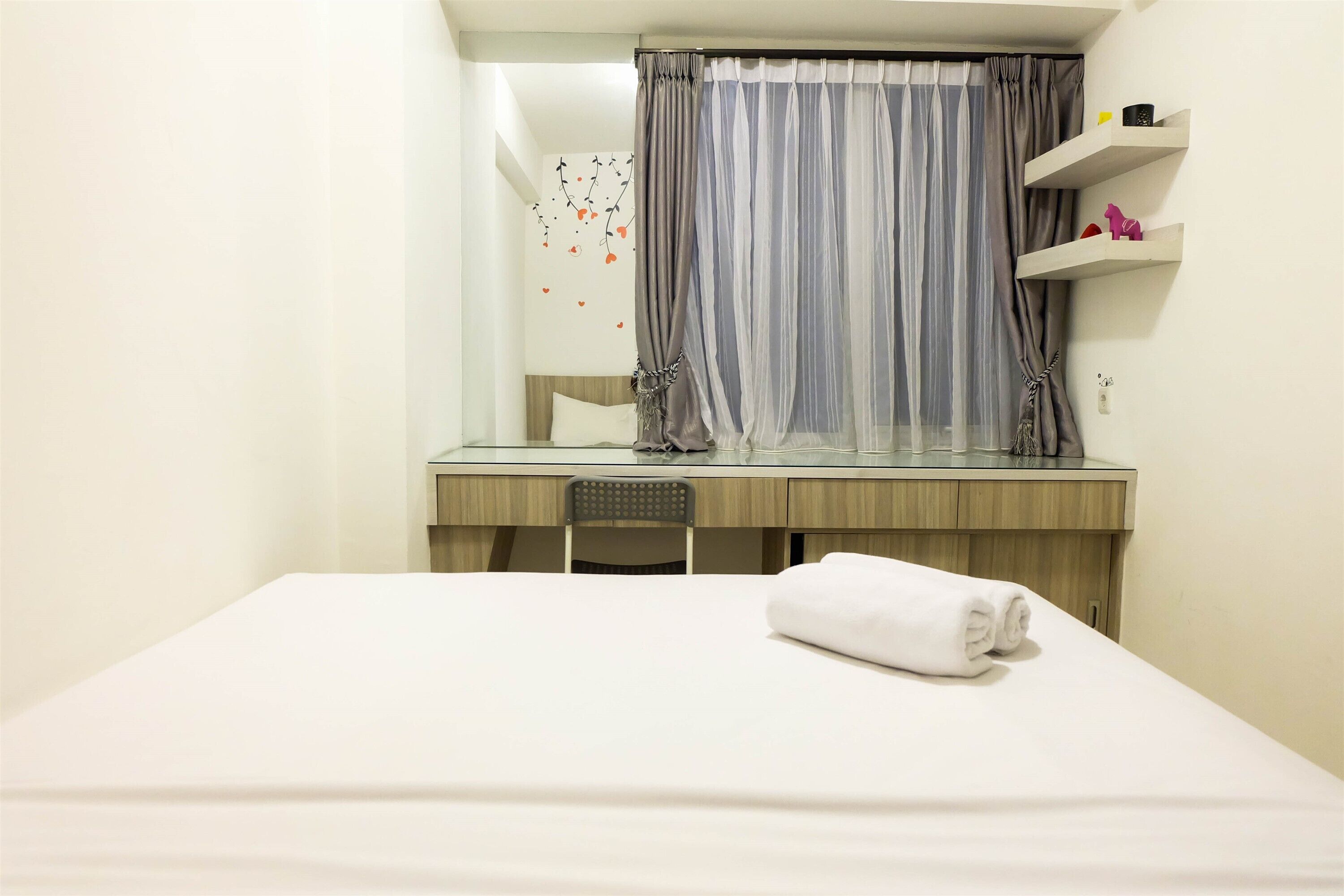 Een hotelkamer met een bed, een bureau en een stoel. Boven het bureau hangt een spiegel, en het raam is afgedekt met een gordijn.