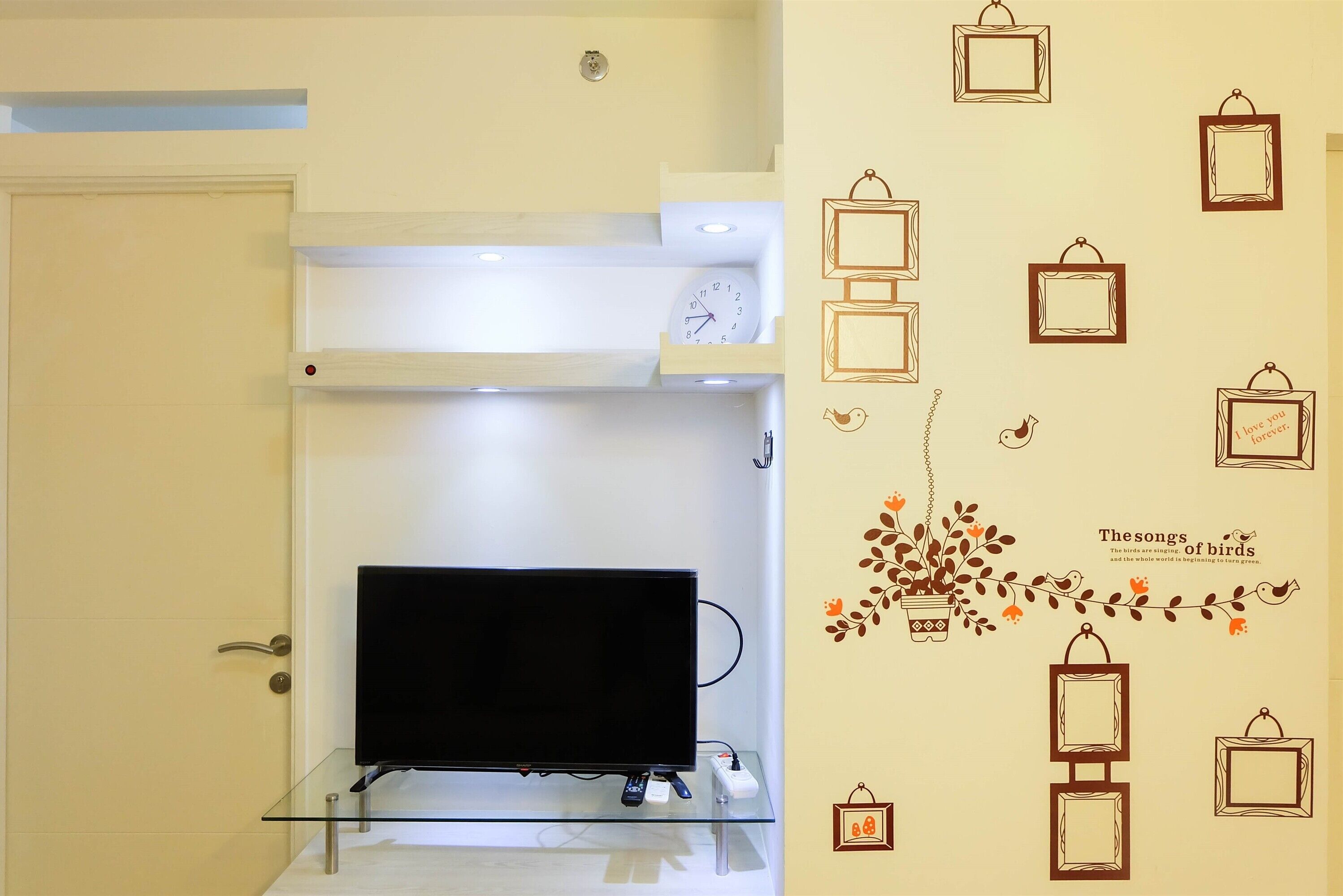 A room with a flat-screen TV on a glass shelf, a door with a handle, and a wall with framed pictures and a decorative bird and plant design.