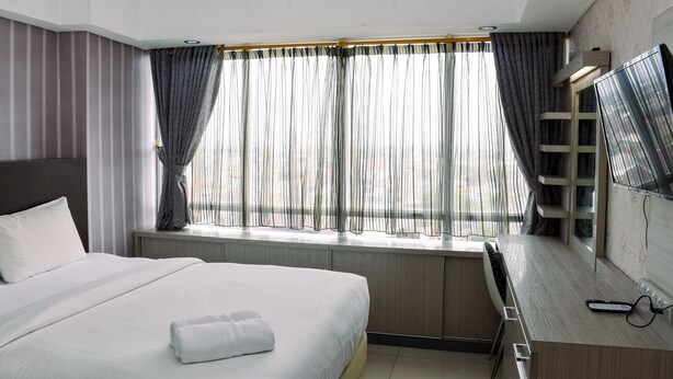 A hotel room with a bed, a desk with a flat-screen TV, a window with curtains, and a wall-mounted TV. - Chic and Cozy 1BR Apartment at H Residence near MT Haryono (Jakarta)