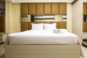 A neatly made bed with white linens and a rolled towel, a large mirror above the bed, and a brown padded headboard.