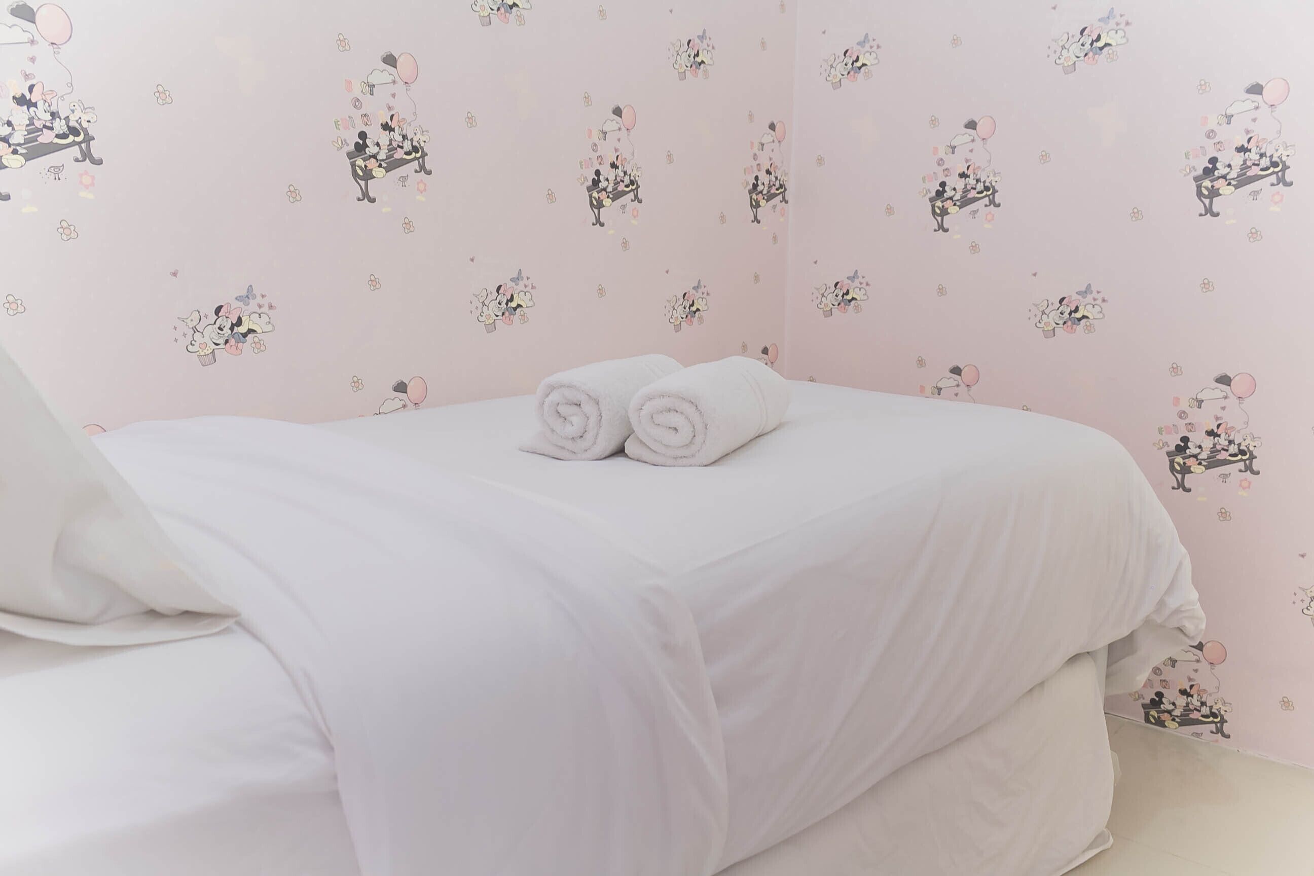 A neatly made bed with white linens and rolled towels, a pink wall with cartoon characters, and a plain white ceiling.