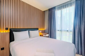 A hotel room with a large bed, a wooden panel wall, a nightstand with a lamp, and a view through sheer curtains.