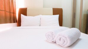 A neatly made bed with white linens and pillows, a rolled white towel, and a wooden headboard.