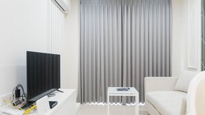 A modern hotel room with a flat-screen TV, a small white table, a sofa, and a large window with grey curtains. - Spacious 1BR Apartment at Praxis (Surabaya)