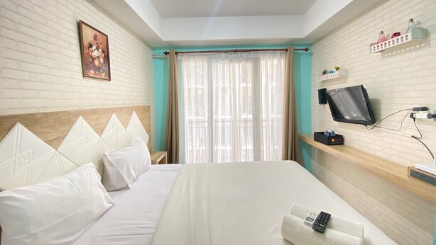 A hotel room with a large bed, a television mounted on the wall, a window with curtains, and a small shelf with decorative items. - Modern and Comfy Studio Room at Grand Asia Afrika Apartment (Bandung)