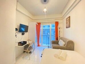 A bedroom with a bed, a desk with a chair, a television, and a balcony with orange curtains. - Cozy Studio Room at Grand Asia Afrika Apartment (Bandung)