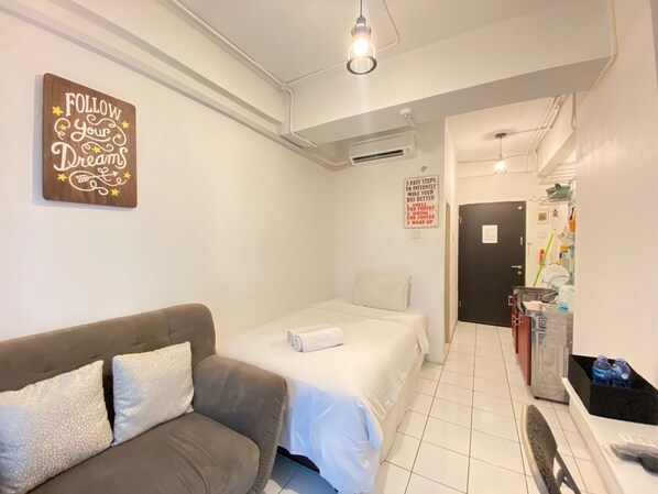 A hotel room with a bed, a sofa, a wall-mounted sign, a door, and a kitchenette area. - Cozy Studio Room at Grand Asia Afrika Apartment (Bandung)