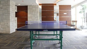 Une table de ping-pong dans une pièce dotée d’un tapis à motifs, de murs en bois et en pierre, et offrant une vue sur des palmiers par la fenêtre.