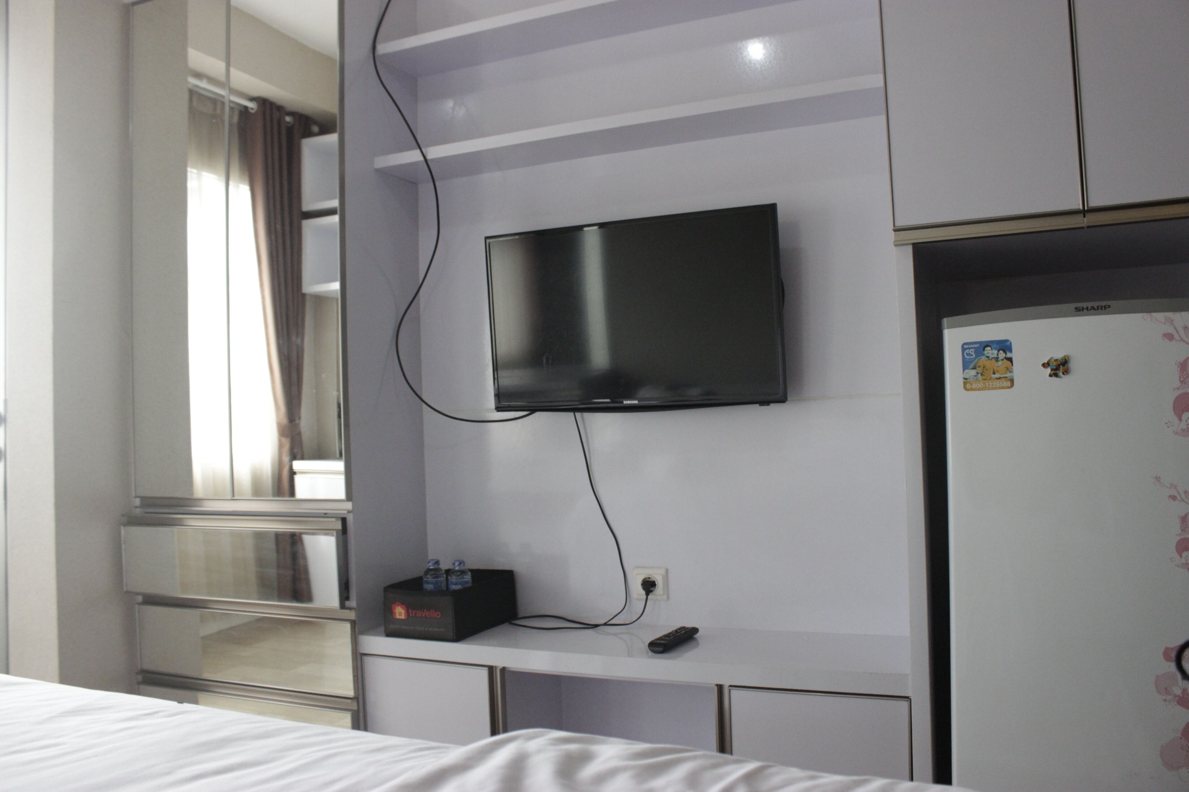 A hotel room with a bed, a flat-screen TV mounted on the wall, a minibar, and a refrigerator with magnets.