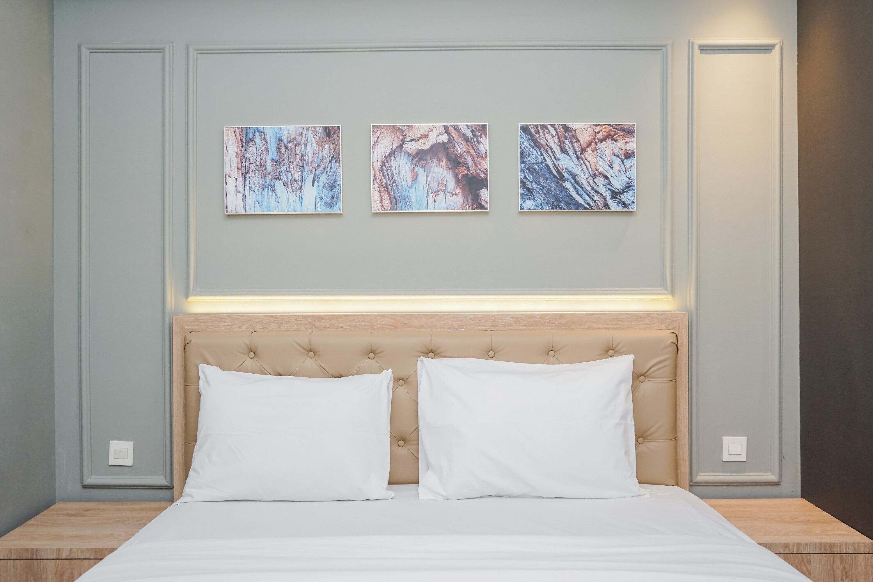 A neatly made bed with two white pillows, a tufted headboard, and three abstract paintings on the wall above.