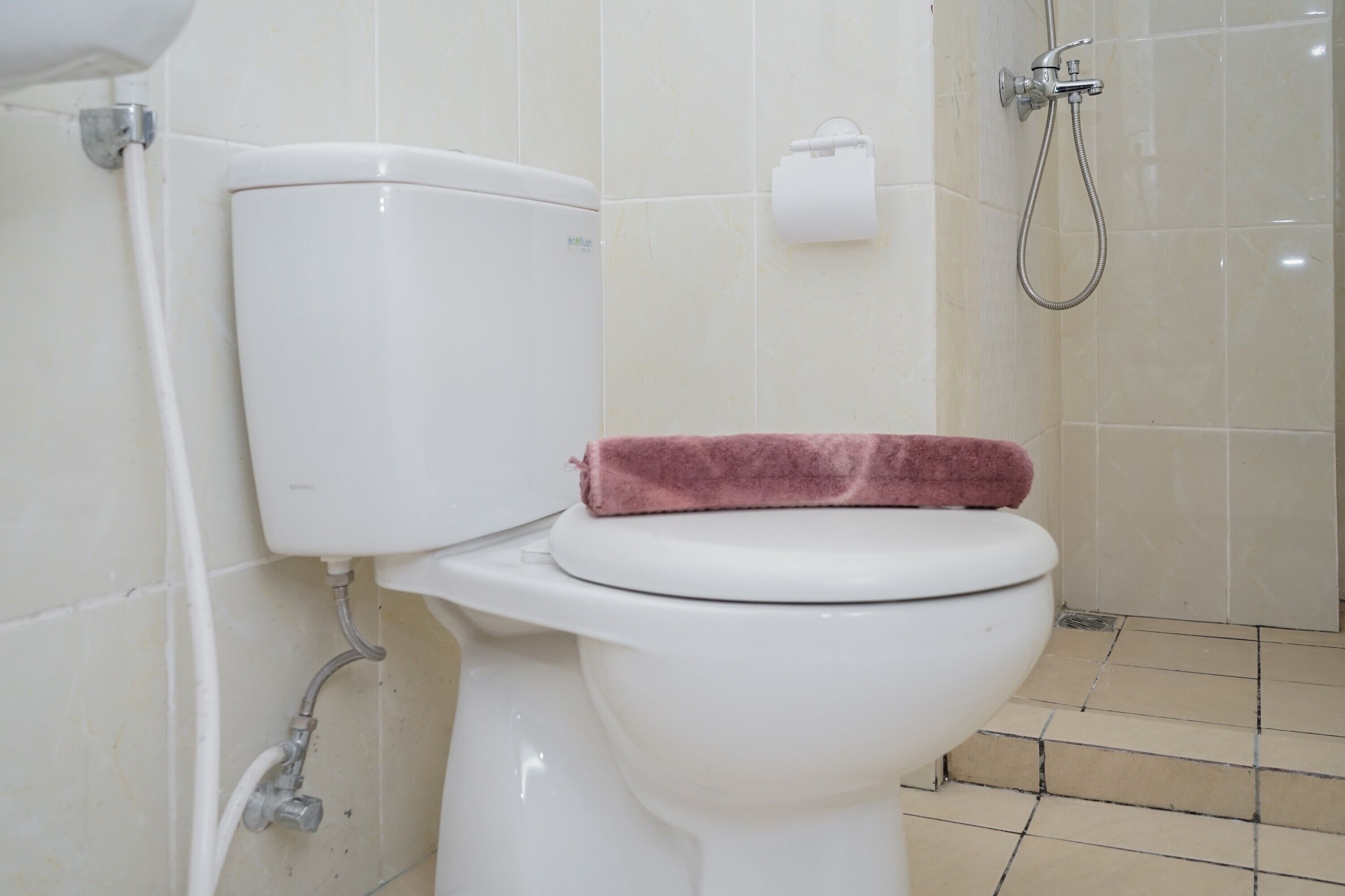 Inodoro blanco con una toalla rosa sobre el asiento, dispensador de papel y zona de ducha con una manguera.