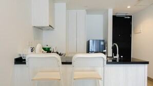 Eine moderne Küche mit weißen Schränken, einer schwarzen Arbeitsplatte und einem Edelstahl-Kühlschrank. Zwei weiße Stühle mit Holzsitzen sowie eine kleine Kücheninsel mit Spüle und Wasserkocher.