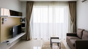 Un salón moderno con un televisor de pantalla plana sobre un estante blanco, un sofá marrón, una mesita pequeña y cortinas translúcidas en un ventanal grande.