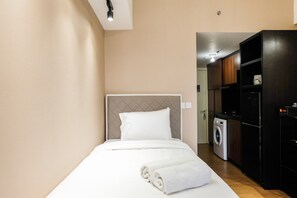 Un lit bien fait, avec un oreiller blanc et une serviette blanche roulée, une machine à laver et un meuble noir dans une petite pièce aux murs de couleur beige.