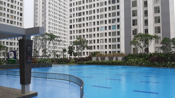 A swimming pool with a curved metal railing, surrounded by lush greenery and tall buildings in the background. - Studio Room Apartment at M-Town Residence near Summarecon Mall Serpong (Gading Serpong)