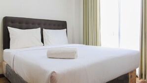 A neatly made bed with white linens and a folded towel, a dark upholstered headboard, and a window with sheer curtains.