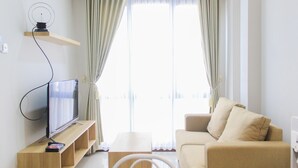 A modern living room with a flat-screen TV on a wooden stand, a beige sofa, and a large window with sheer curtains.