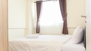 A neatly made bed with a white comforter and pillows, a window with brown curtains, and a wooden door frame. - Spacious and Well Appointed 2BR at Meikarta Apartment (Cikarang)