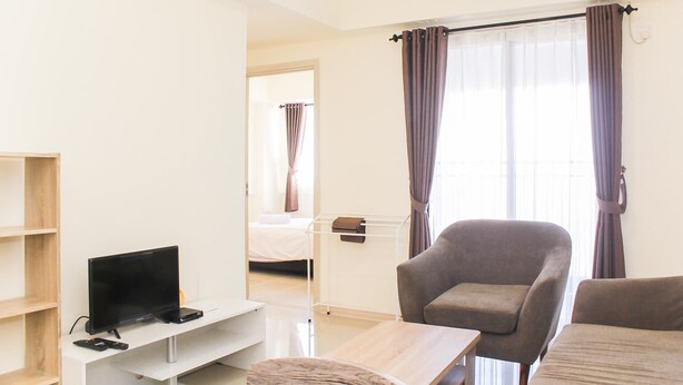 A hotel room with a TV, a chair, a small table, and a bed in the background. - Spacious and Well Appointed 2BR at Meikarta Apartment (Cikarang)