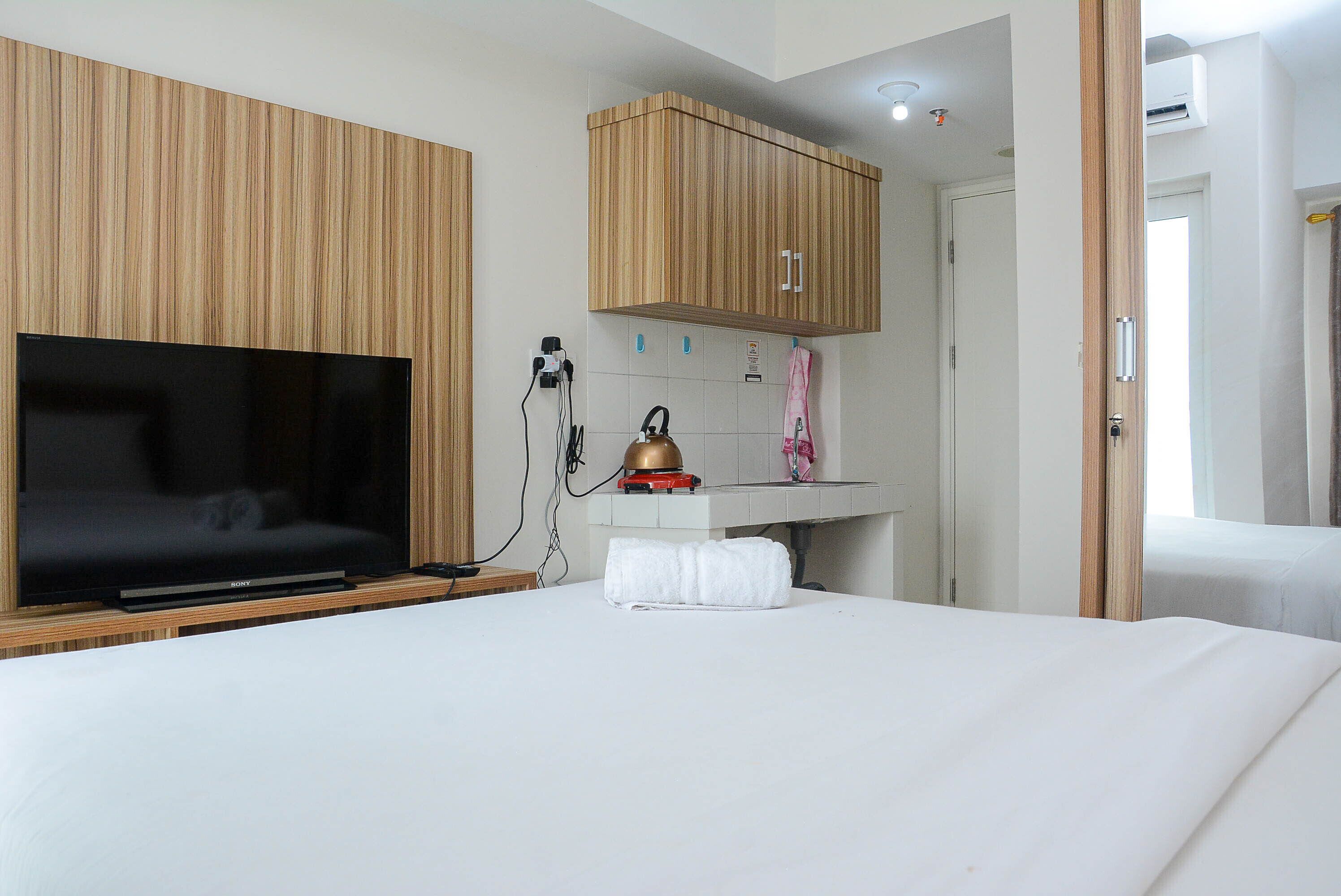 A hotel room with a large flat-screen TV, wooden paneling, a small kitchen area with a kettle, and a neatly made bed.