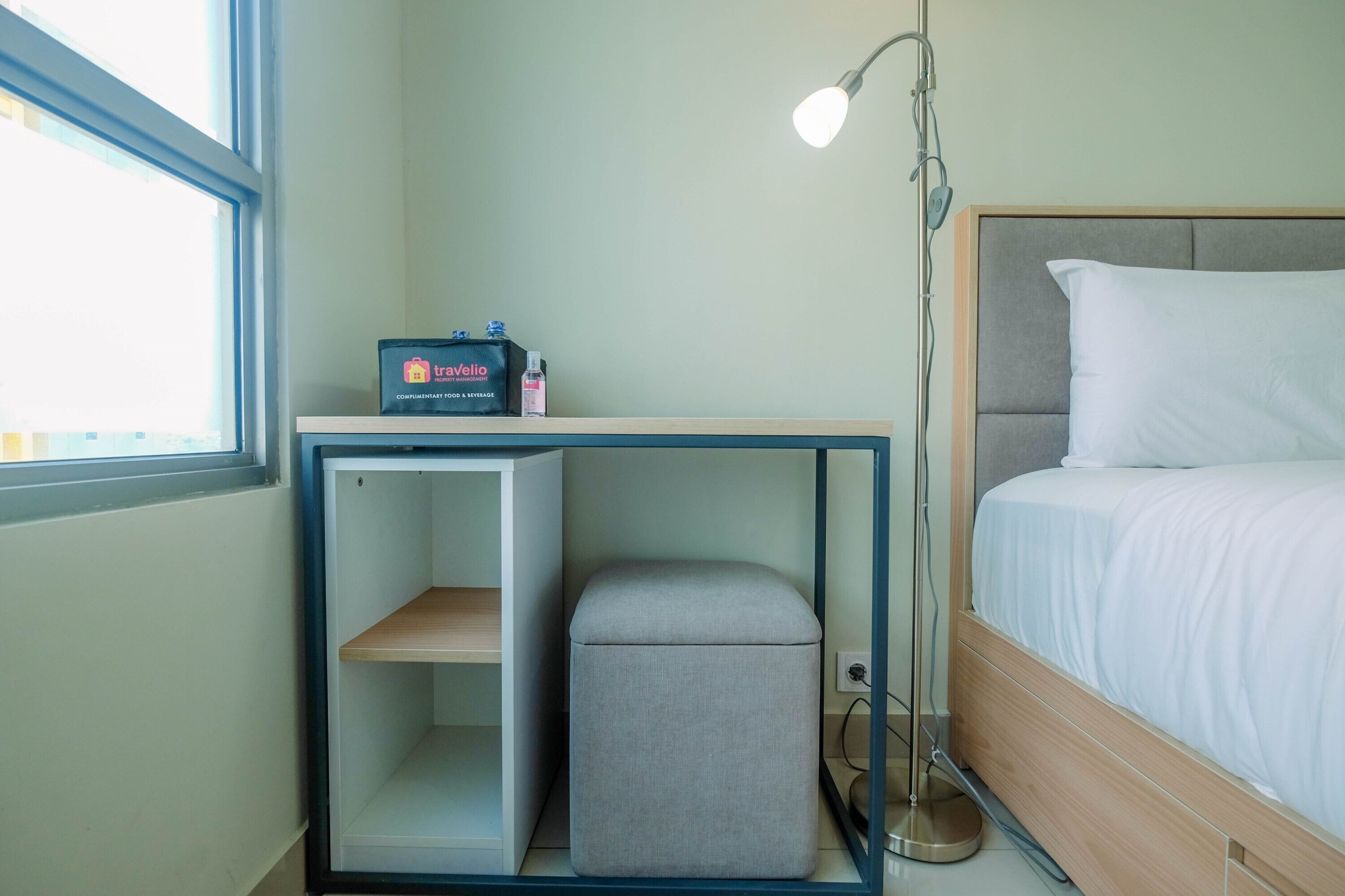 A hotel room with a bed, a bedside table with a lamp, a small stool, and a shelf with toiletries.
