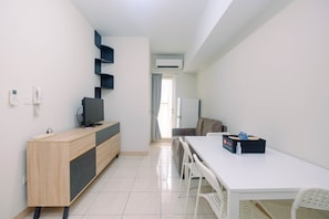 Une pièce moderne et bien éclairée, avec un meuble en bois, un téléviseur à écran plat, une table à manger blanche avec des chaises et un réfrigérateur.