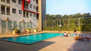 A swimming pool area with lounge chairs and a building with multiple windows and balconies. - Cozy Studio at Elvis Tower Apartment (Cikarang)