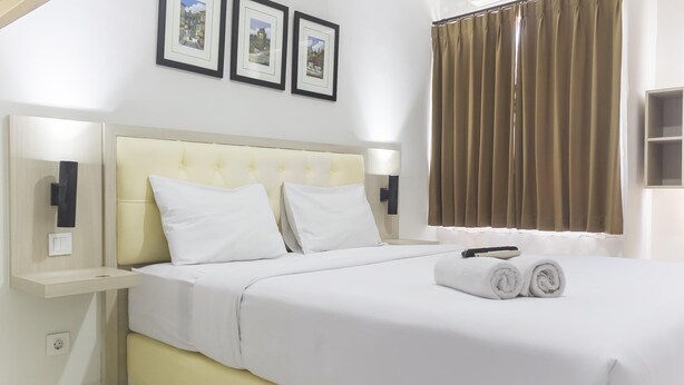 A neatly made bed with white linens and two pillows, a bedside table with a lamp, and a window with brown curtains. - Cozy Studio at Elvis Tower Apartment (Cikarang)