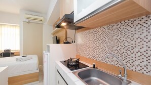 Eine kleine Küchenzeile mit Gasherd, Spüle und Mikrowelle. Ein Bett mit weißer Matratze sowie ein kleiner Schreibtisch mit Stuhl sind ebenfalls vorhanden.