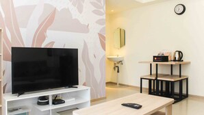 Ett modernt vardagsrum med en platt-TV på ett vitt TV-bord, ett träbord som används som soffbord, ett litet bord med en vattenkokare och koppar samt en väggklocka.