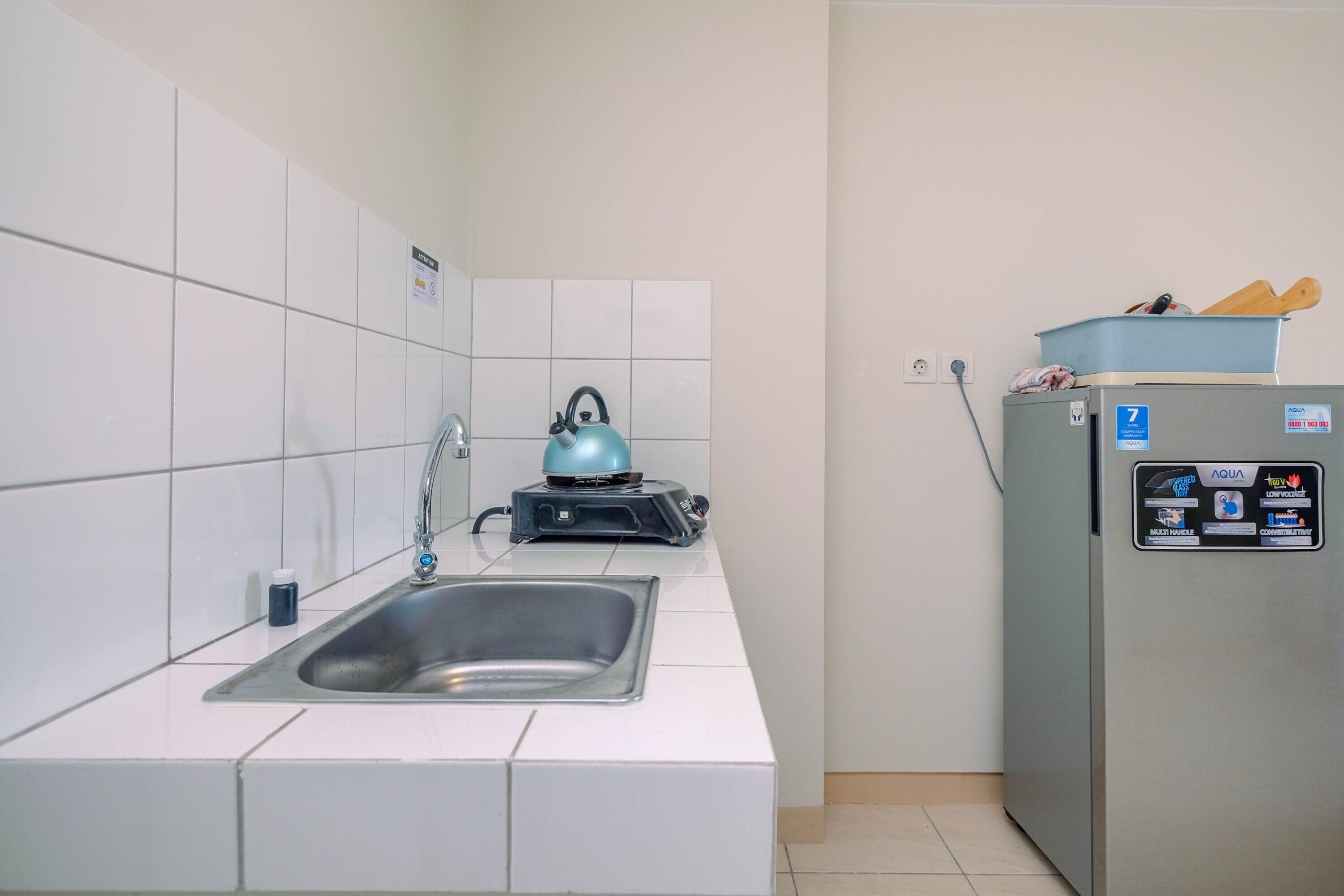 A small kitchen with a stainless steel sink, a kettle, and a refrigerator.