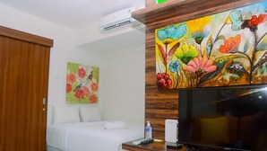 A hotel room with a bed, a television, a painting, and an air conditioning unit. - Cozy and Simple Living 1BR Grand Kamala Lagoon Apartment (Bekasi)