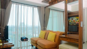 A modern living room with a sofa, a small table, a television, and a large window with curtains. - Cozy and Simple Living 1BR Grand Kamala Lagoon Apartment (Bekasi)