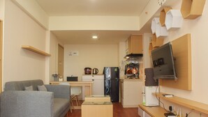 A compact living space with a grey sofa, a small wooden coffee table, a kitchen area with a refrigerator, stove, and microwave, and a mounted TV. - Comfort and Nice 2BR at Meikarta Apartment (Cikarang)