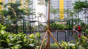 Uma área de ginástica ao ar livre com equipamentos de exercício, rodeada por vegetação exuberante e edifícios altos ao fundo.