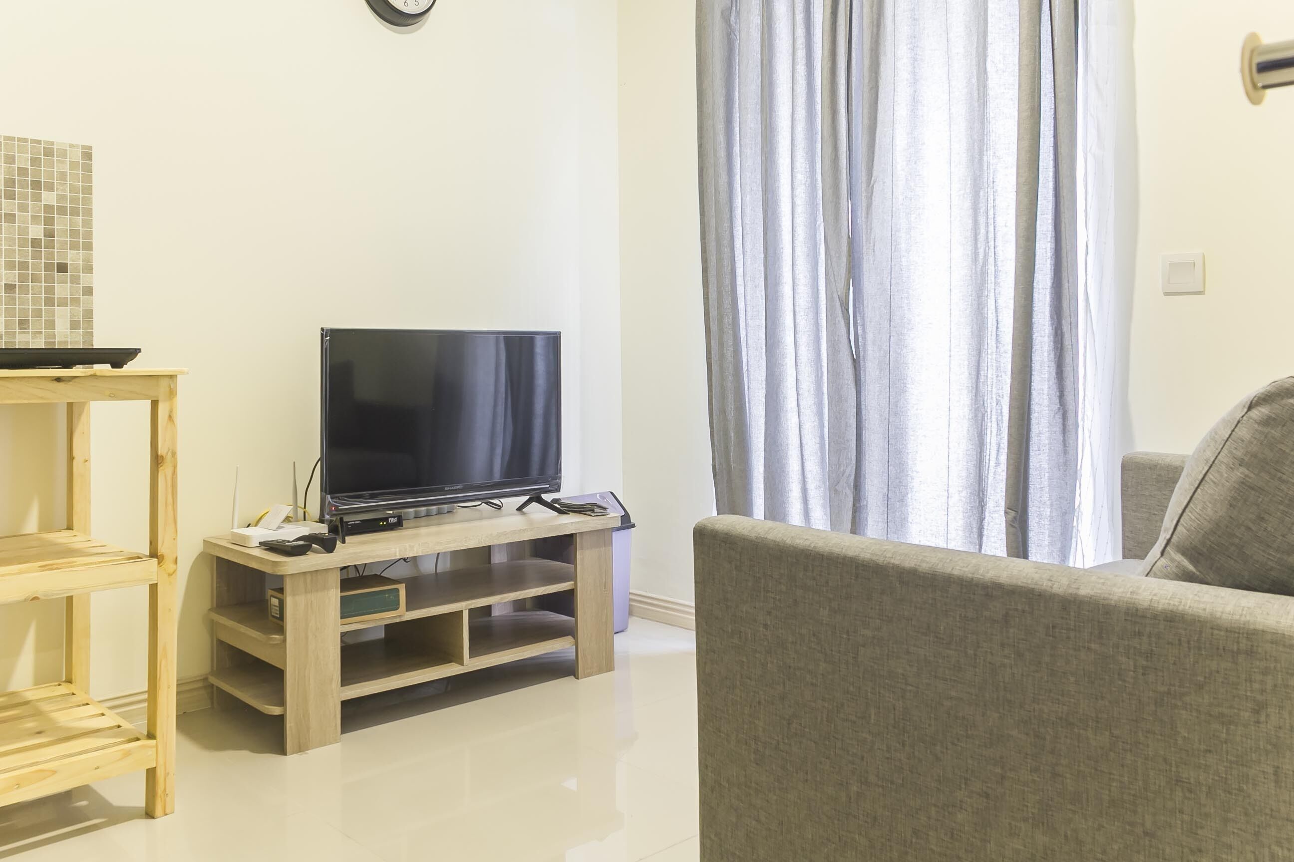 A living room with a TV on a wooden stand, a grey sofa, and a window with curtains.