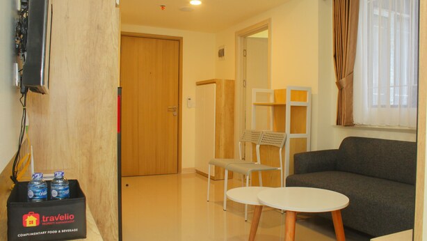 A living room with a sofa, a small round table, and a chair. There is a wooden door, a white chair, and a shelf with a mirror. A box labeled 'Travelio' is on the floor. - Cozy and Relax 2BR at Meikarta Apartment (Cikarang)