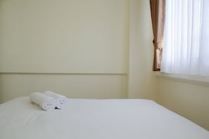 A neatly made bed with two rolled towels on it, a plain wall, and a window with curtains.