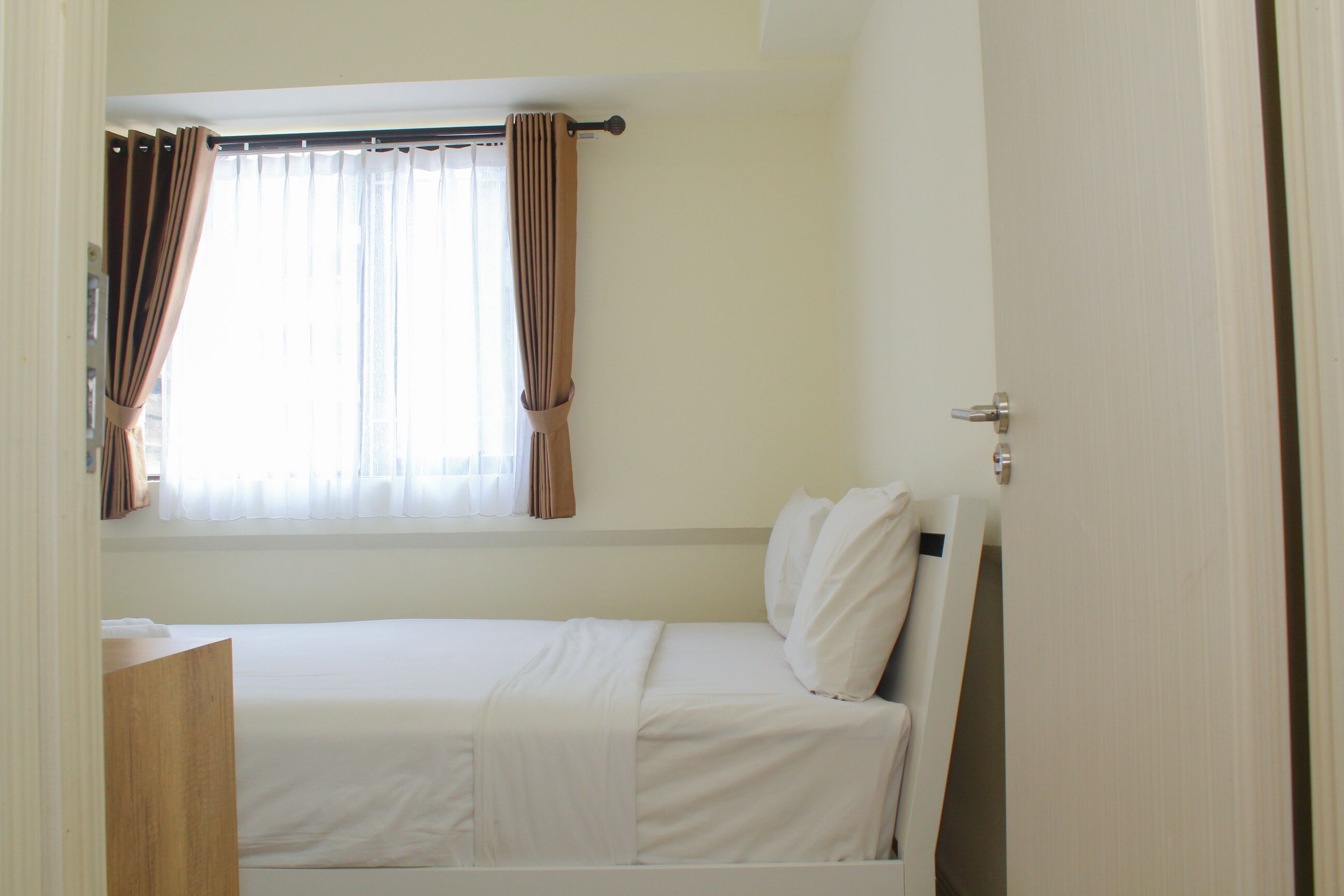Una cama individual con ropa blanca, una ventana con cortinas y una mesita de noche de madera.