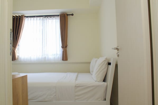 A single bed with white bedding, a window with curtains, and a wooden nightstand.